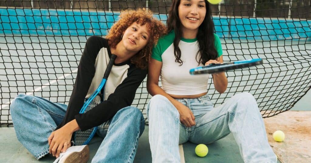 girls with tennis rackets bouncing tennis balls on their racket, sitting in front of a net